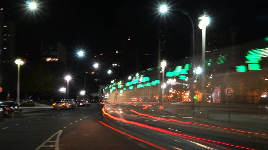 Nighttime city street scene with bright streetlights, blurred car light trails on the road, and a moving train with green illuminated signs creating a streaked light effect. Buildings are visible in the background.