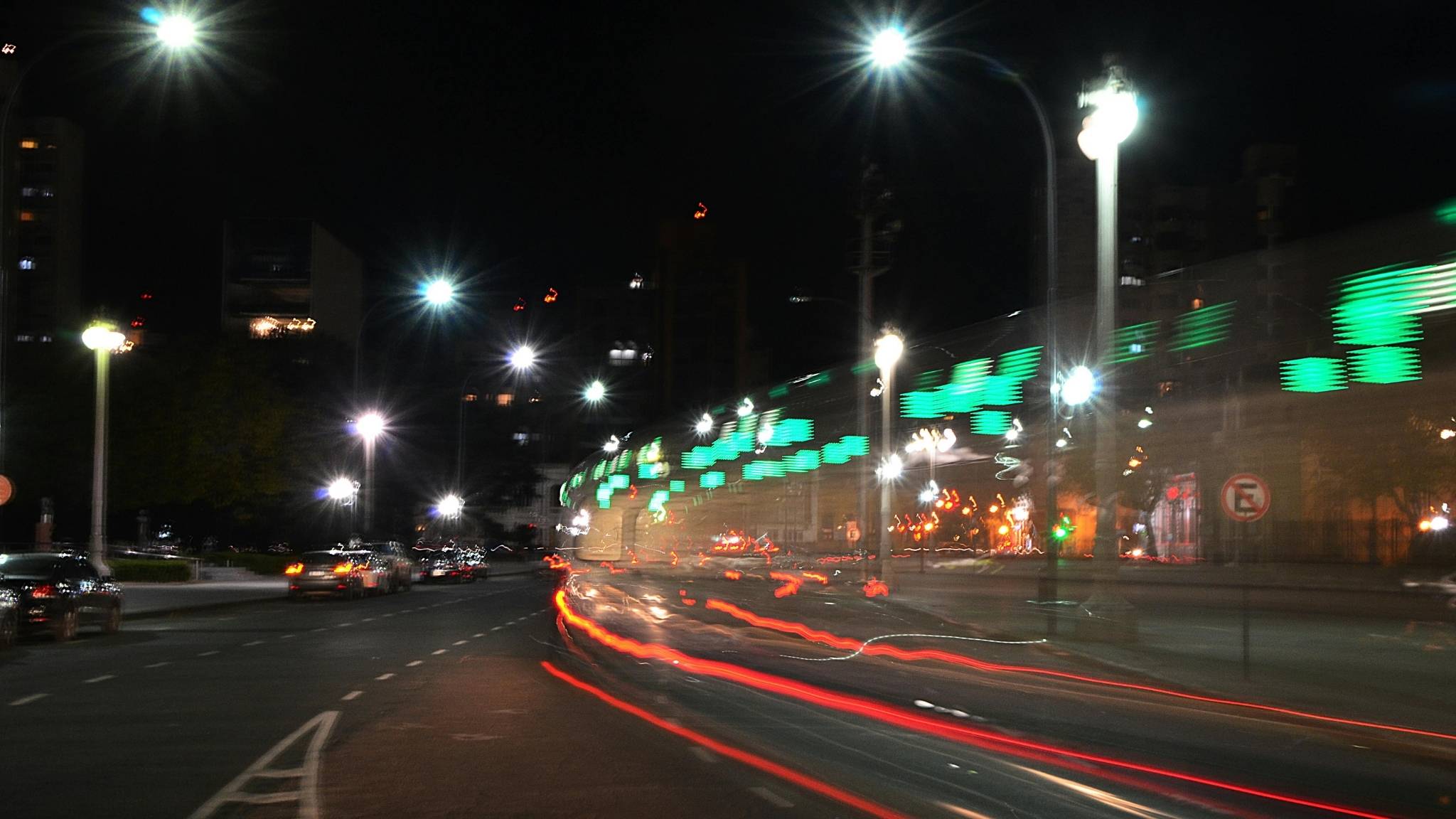 Nighttime city street scene with bright streetlights, blurred car light trails on the road, and a moving train with green illuminated signs creating a streaked light effect. Buildings are visible in the background.