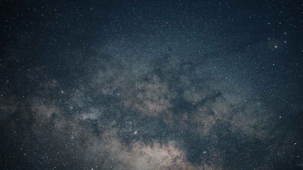 Night sky filled with countless stars and galaxies, with a faint shooting star or meteor streaking across a section of the image. The Milky Way is clearly visible, creating a cloudy band of light and dark patches.