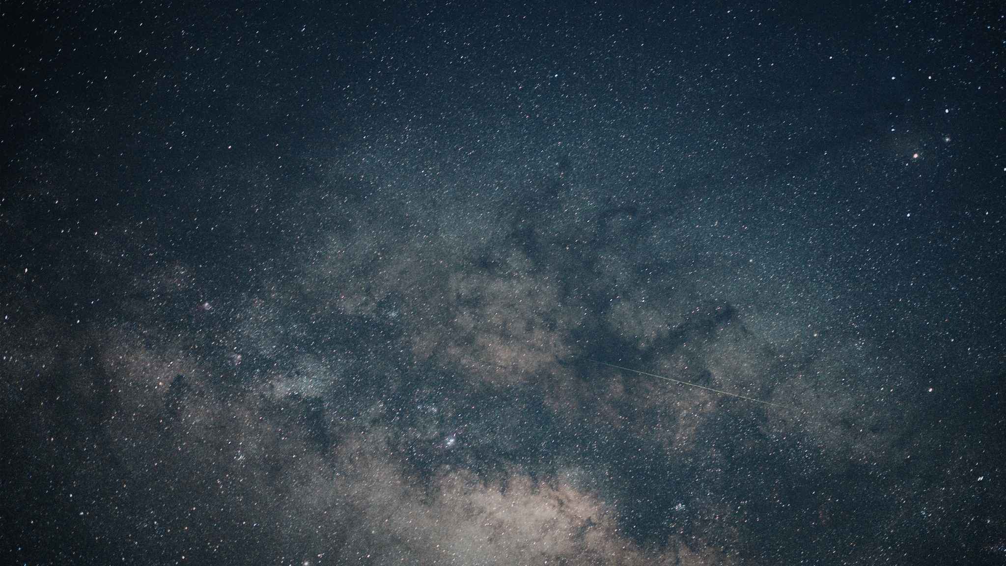 Night sky filled with countless stars and galaxies, with a faint shooting star or meteor streaking across a section of the image. The Milky Way is clearly visible, creating a cloudy band of light and dark patches.