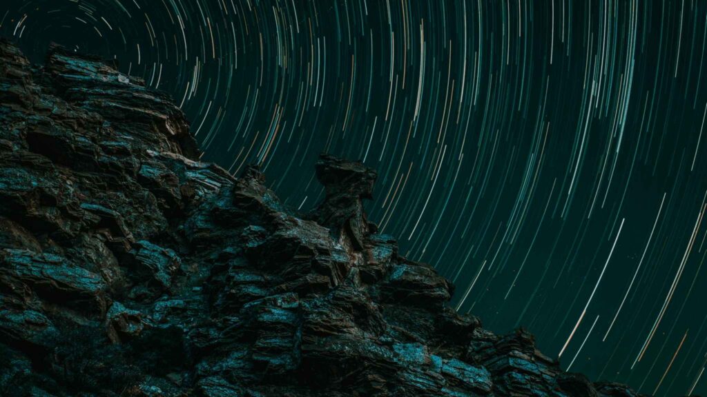 Jagged rock formation in the foreground with circular star trails in the night sky above, showing the motion of stars due to long-exposure photography.