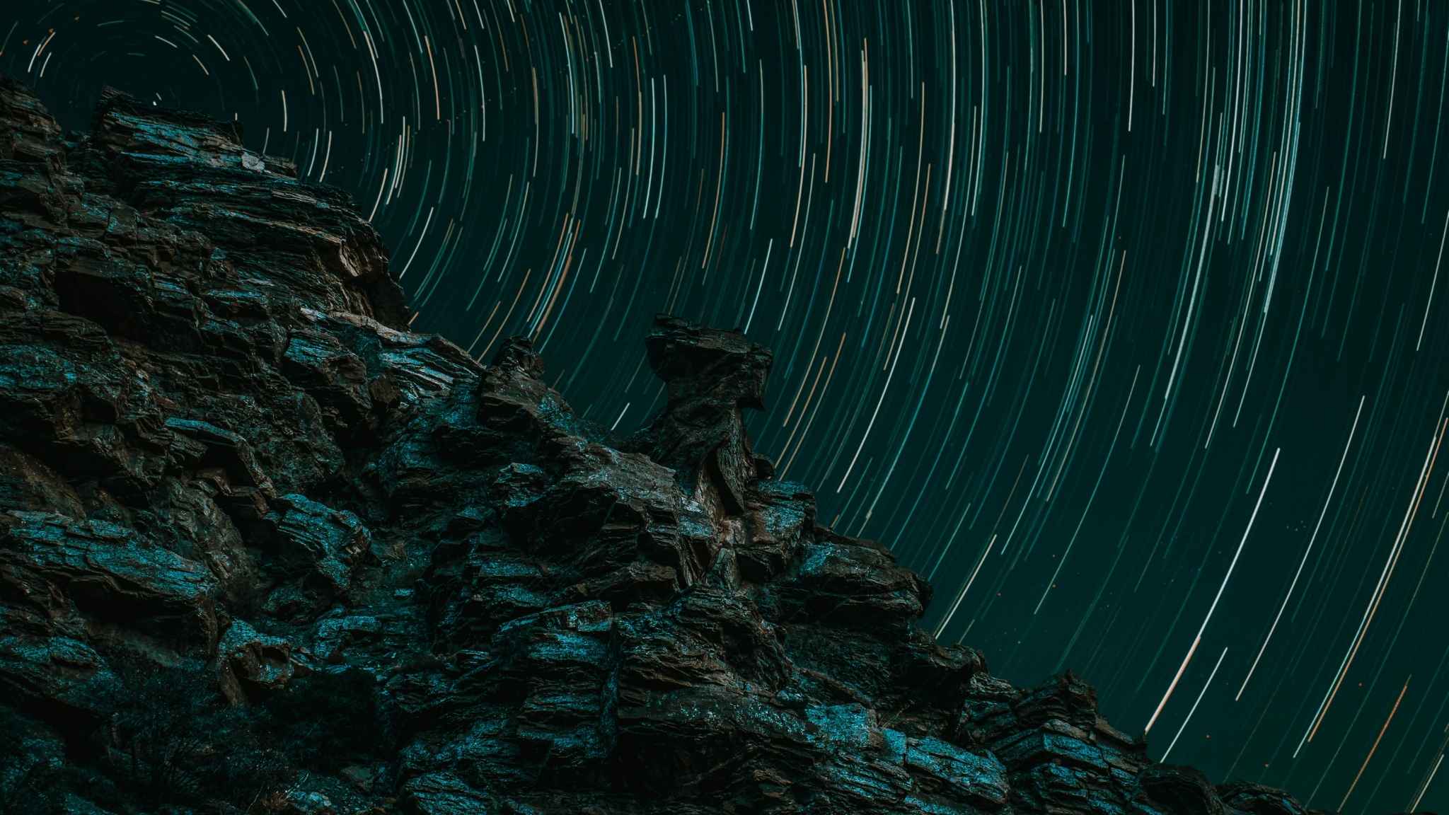 Jagged rock formation in the foreground with circular star trails in the night sky above, showing the motion of stars due to long-exposure photography.