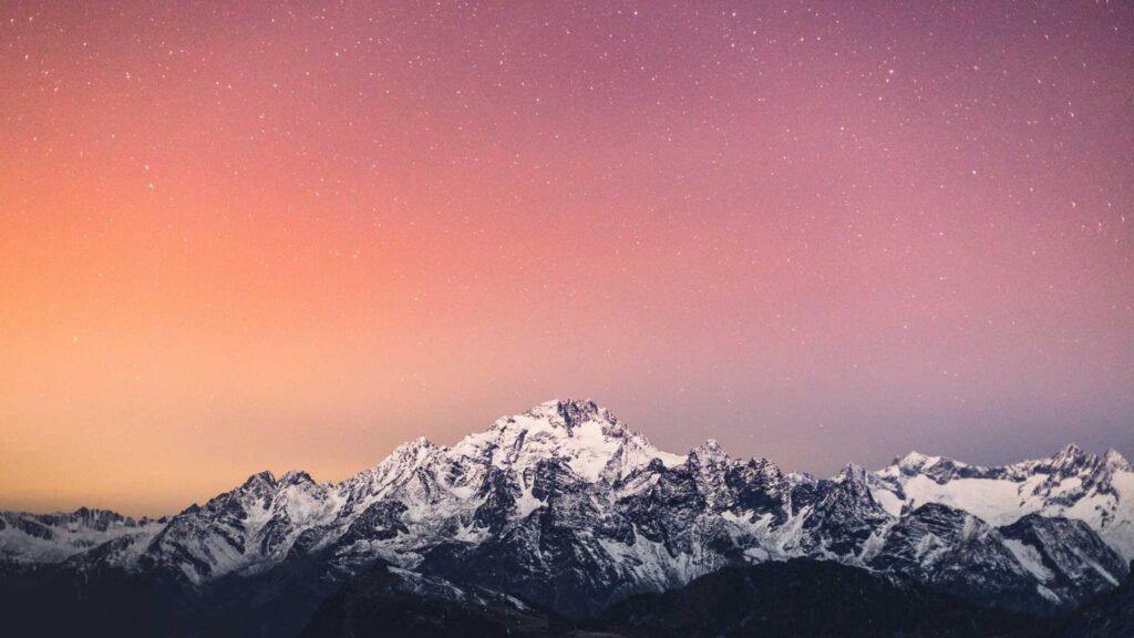 Snowy mountain range under a vibrant, colorful sky at dusk, with stars beginning to appear above the peaks.
