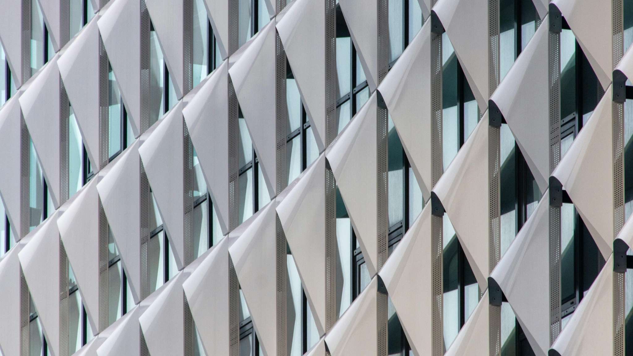 Close-up of a modern building facade with diagonal, overlapping white panels and glass windows, creating a geometric, repetitive pattern.