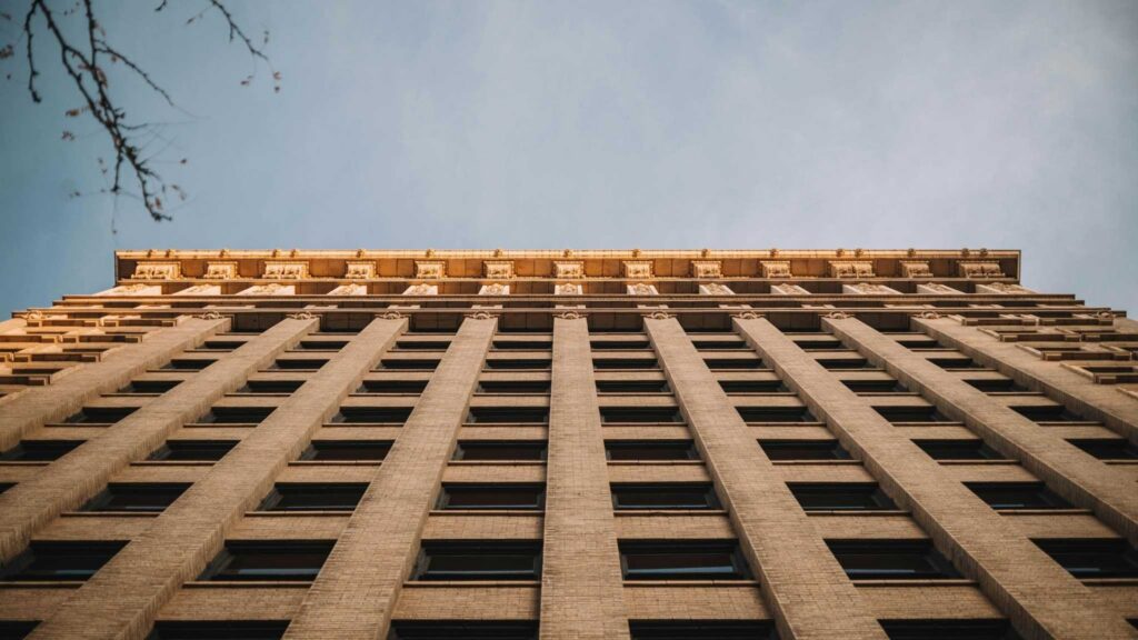 Tall beige brick building shot from below, with multiple windows in vertical rows, bathed in warm sunlight at the top, and a clear sky in the background. Branches appear in the upper left corner.