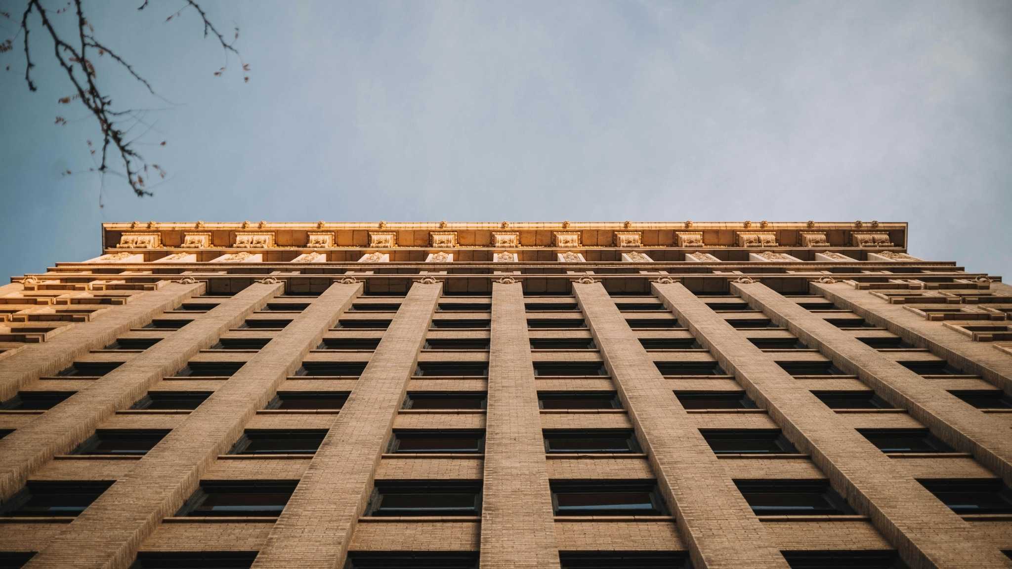 Tall beige brick building shot from below, with multiple windows in vertical rows, bathed in warm sunlight at the top, and a clear sky in the background. Branches appear in the upper left corner.