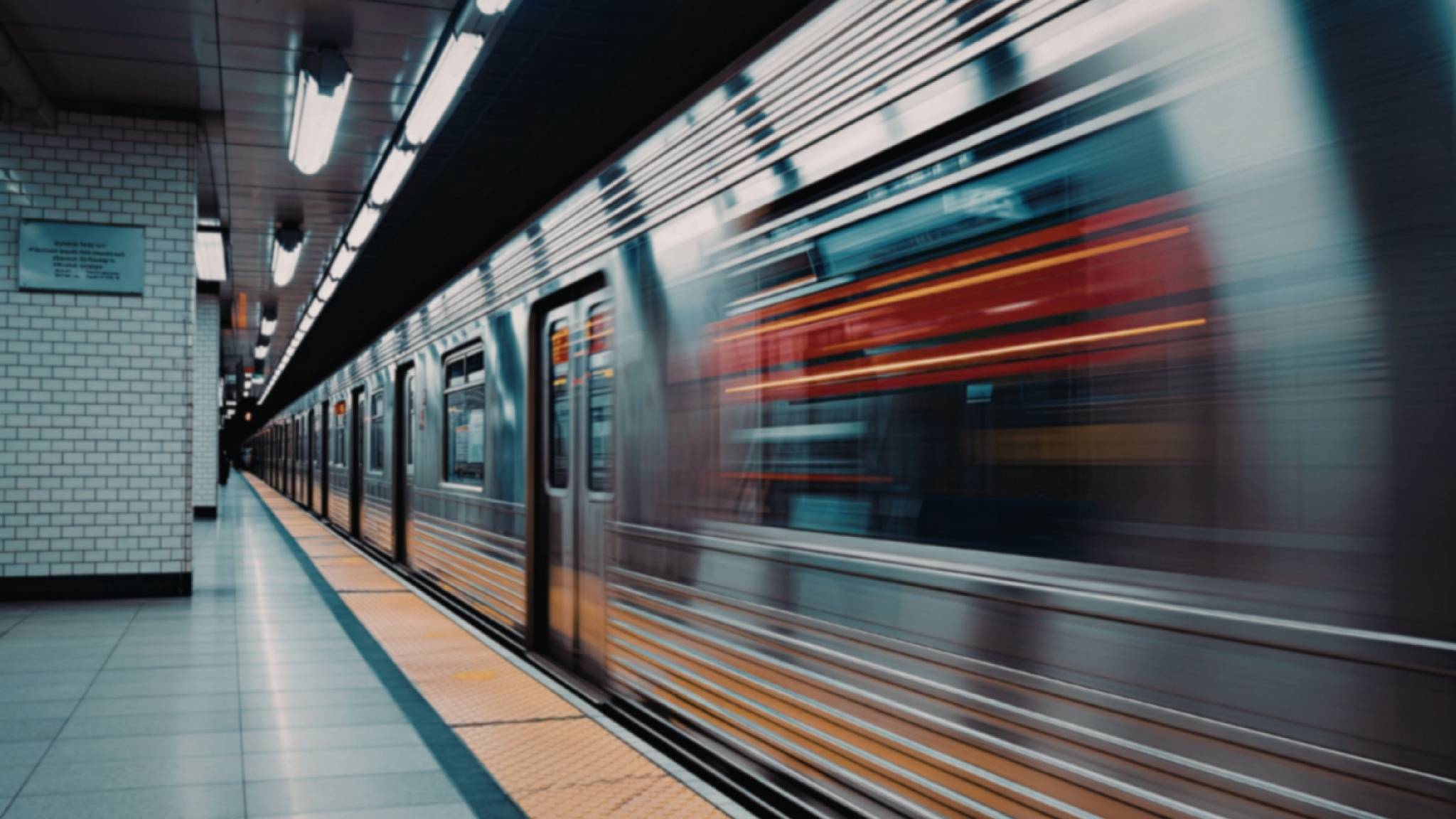 A subway train speeds past a tiled underground station platform, creating a motion blur effect. The platform is empty, and fluorescent lights illuminate the scene.