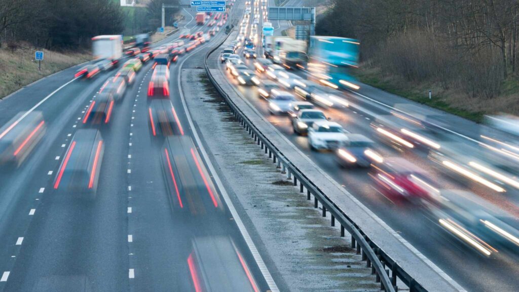 Cars and trucks drive fast on a busy multi-lane highway, captured with motion blur. Traffic is heavier on the right side of the road, while the left side has fewer vehicles. Trees and road signs are visible in the background.