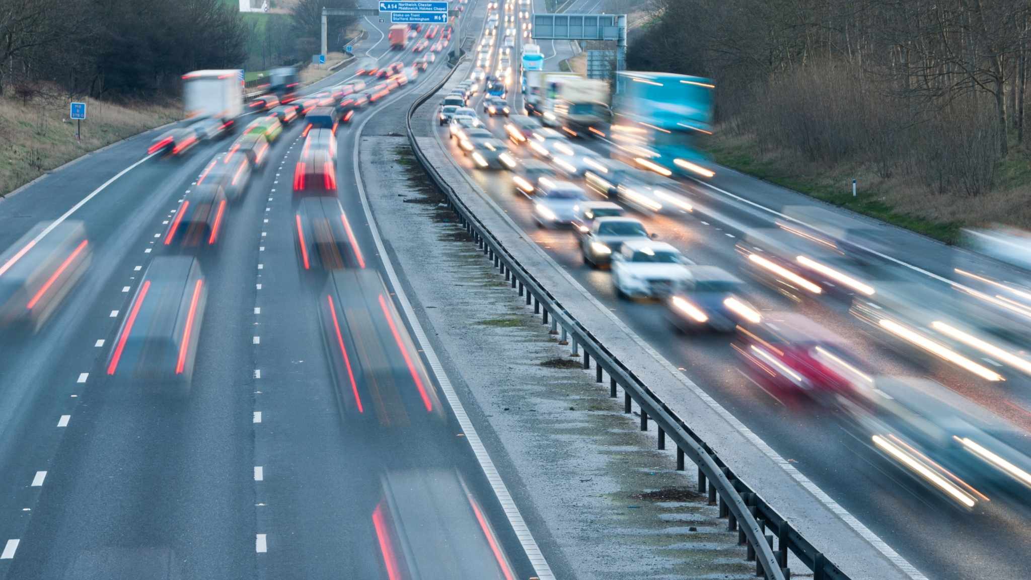 Cars and trucks drive fast on a busy multi-lane highway, captured with motion blur. Traffic is heavier on the right side of the road, while the left side has fewer vehicles. Trees and road signs are visible in the background.