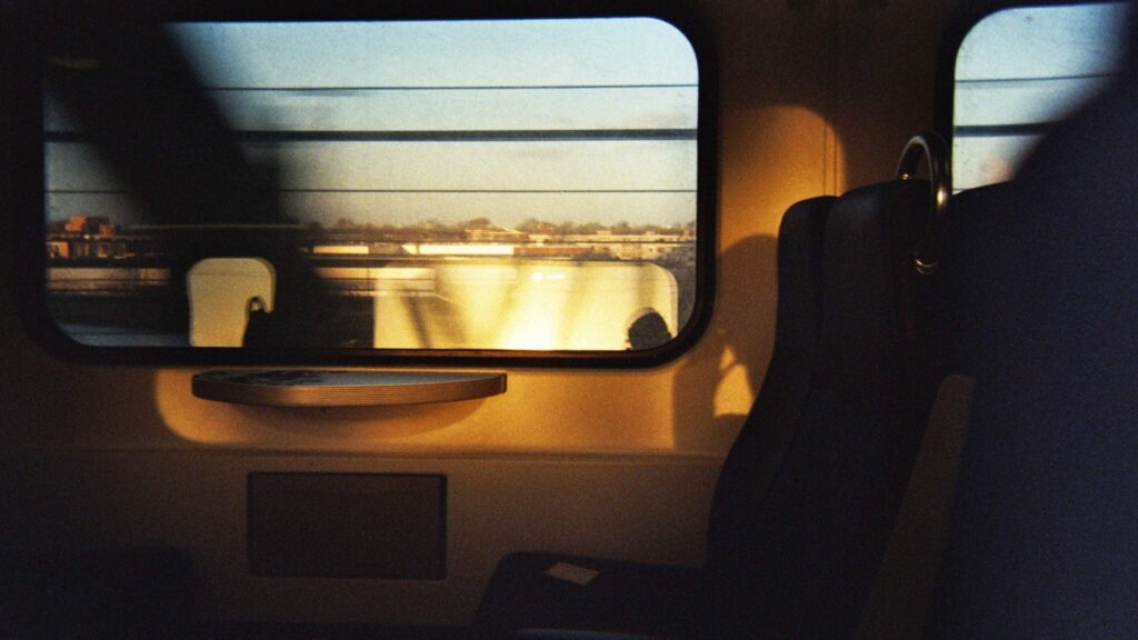 Sunlight shines through a train window onto an empty seat and table, casting shadows. Outside, blurred buildings and a fence are visible, suggesting the train is in motion. The scene feels calm and quiet.