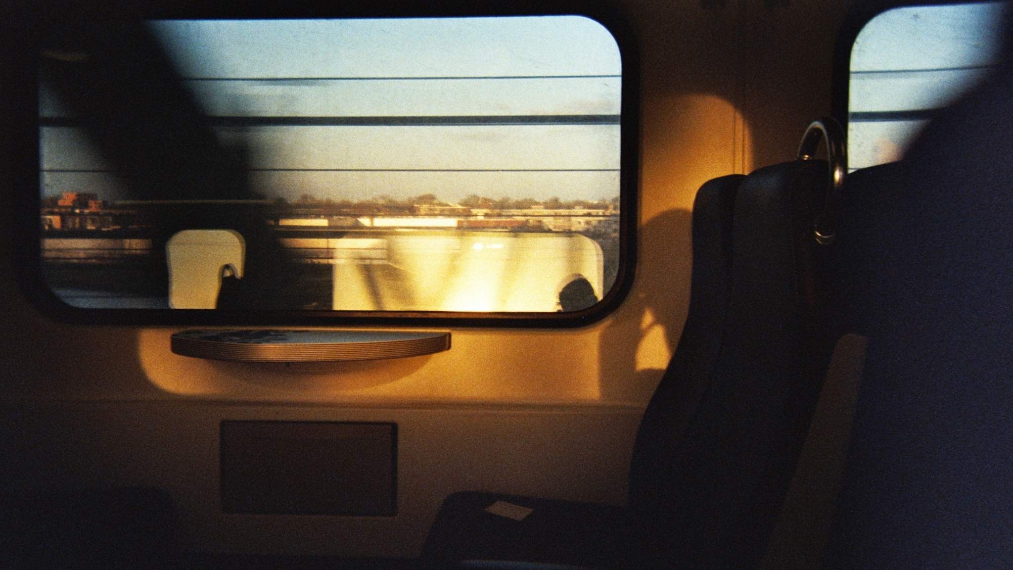 Sunlight shines through a train window onto an empty seat and table, casting shadows. Outside, blurred buildings and a fence are visible, suggesting the train is in motion. The scene feels calm and quiet.