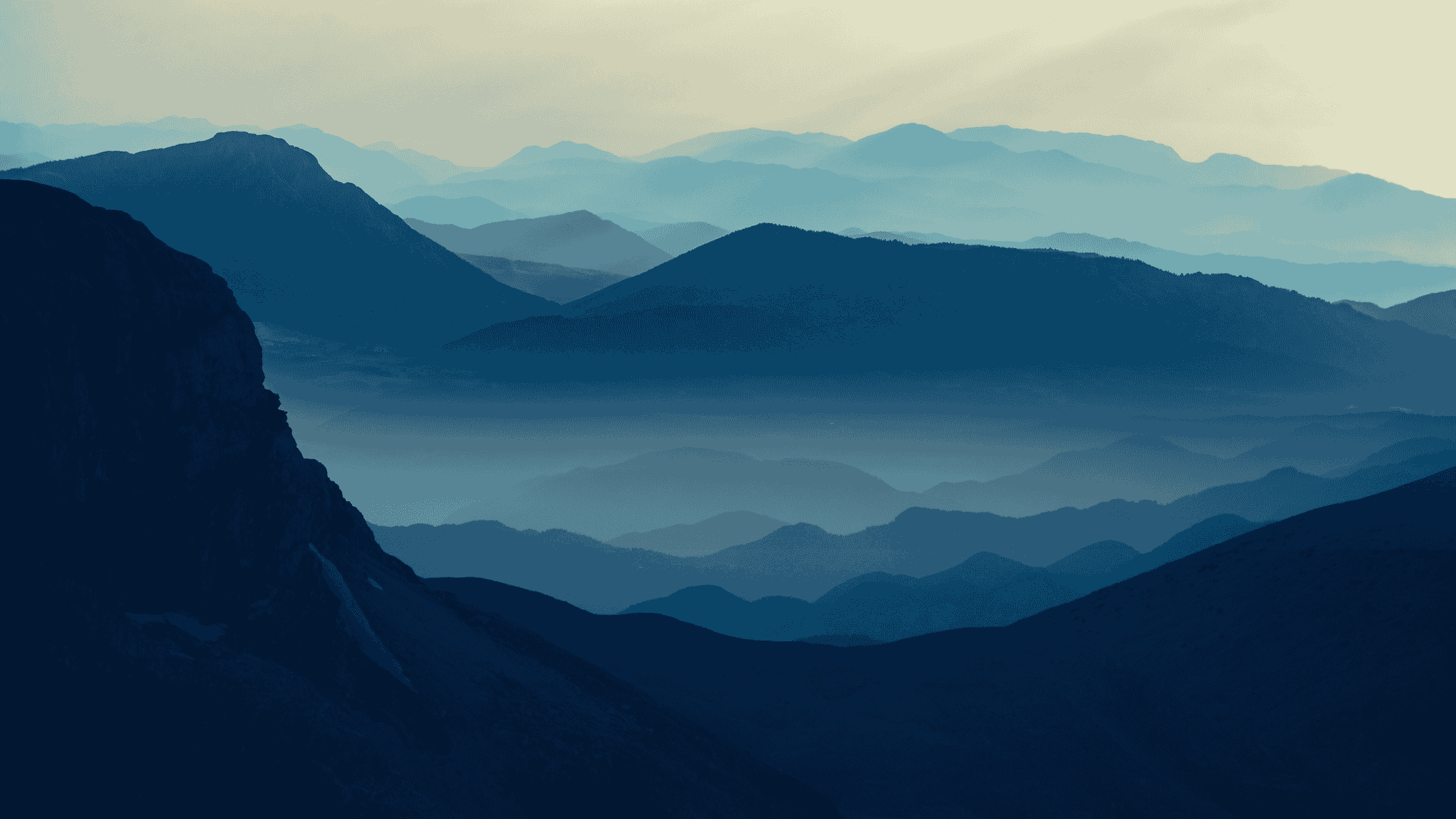Blue-toned silhouettes of mountain ranges overlap under a hazy sky, with layers fading into the distance and soft mist filling the valleys, creating a peaceful, atmospheric landscape.