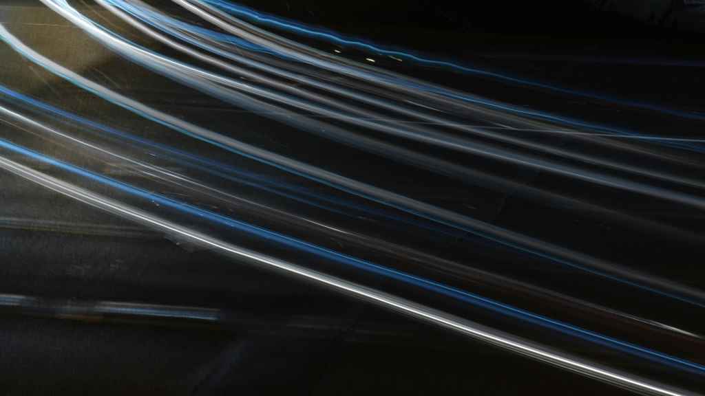 Long white and blue light trails streak diagonally across a dark background, creating an abstract, dynamic effect suggestive of motion or fast-moving vehicles at night.