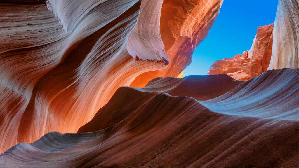 Curved, smooth sandstone walls in shades of orange and blue create a dramatic canyon landscape, with a bright blue sky visible above.