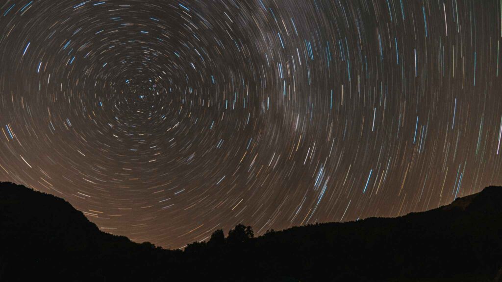 Long exposure photo of a night sky showing circular star trails above dark mountain silhouettes, highlighting the rotation of Earth.
