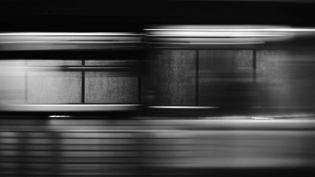 A blurred, black and white image captures the motion of a train passing by a station platform, with horizontal lines emphasizing speed and movement.