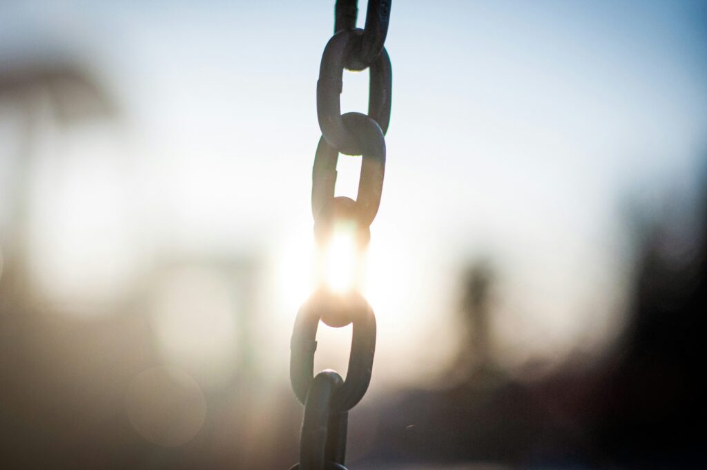 Link integrity checker. A close-up of a metal chain with sunlight shining through the links, creating a bright, blurred background.