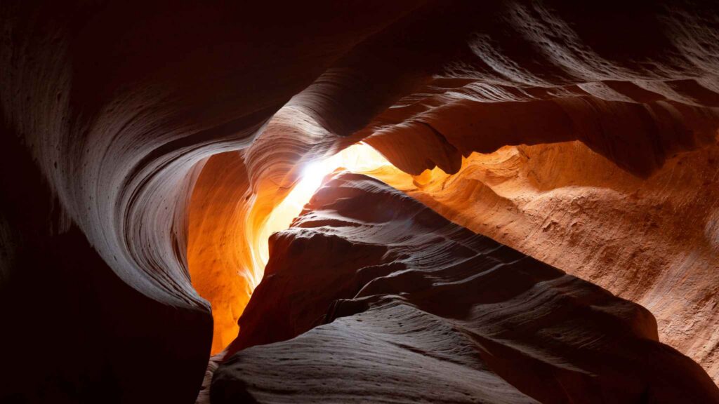 Sunlight streams through narrow, winding sandstone walls in a slot canyon, highlighting smooth, wavy rock textures in shades of orange and brown.