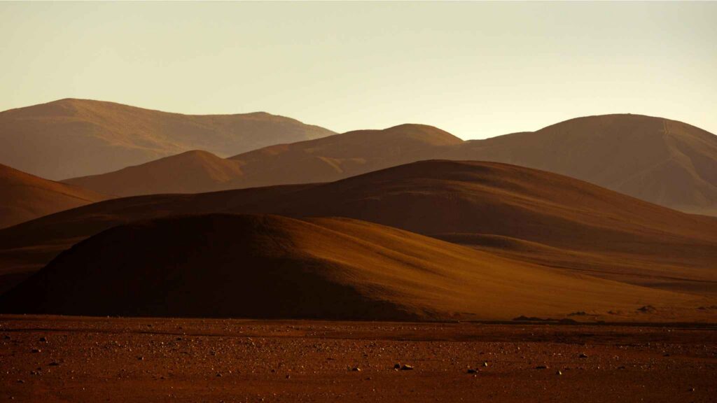 Rolling brown hills under a hazy sky, with soft sunlight highlighting the curves of the landscape and distant mountain ridges in the background.