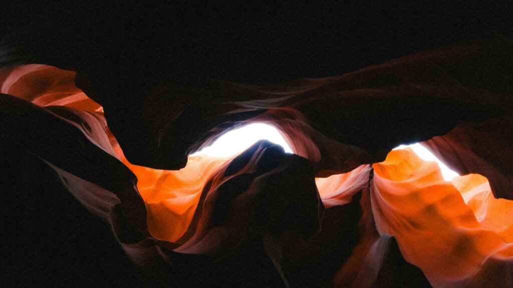 Sunlight shines through the curving, layered walls of a narrow slot canyon, illuminating the reddish-orange rock formations against the dark interior.