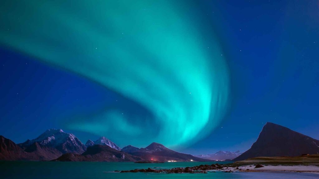 Green northern lights illuminate a clear night sky over snowy mountains and calm water, with a small cluster of lights visible near the shoreline.