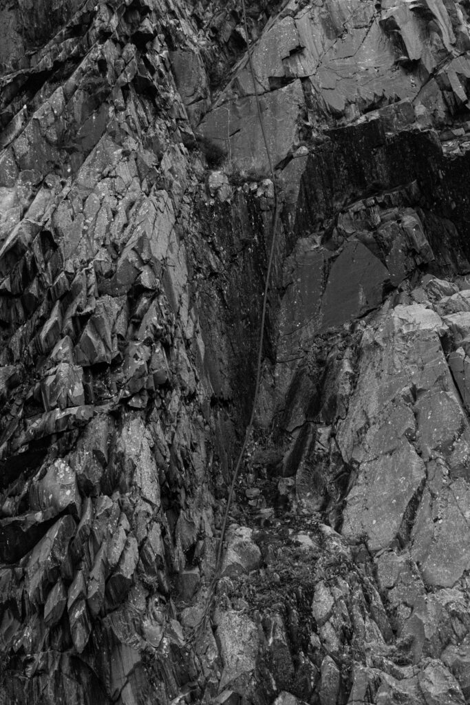 Black-and-white photo of jagged rock formations with sharp edges and layered surfaces. A thin, weathered rope runs diagonally across the middle of the rocks for technical documentation quality.