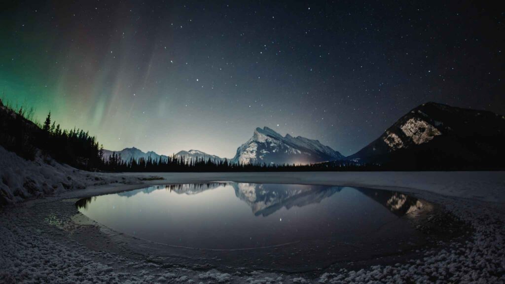 Snowy mountains and a forest are reflected in a calm, partially frozen lake under a starry night sky with faint green northern lights.