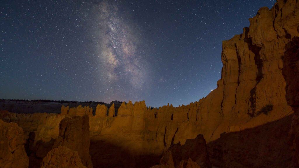 Rocky canyon cliffs under a clear night sky with the Milky Way galaxy visible above, illuminated by soft natural light.