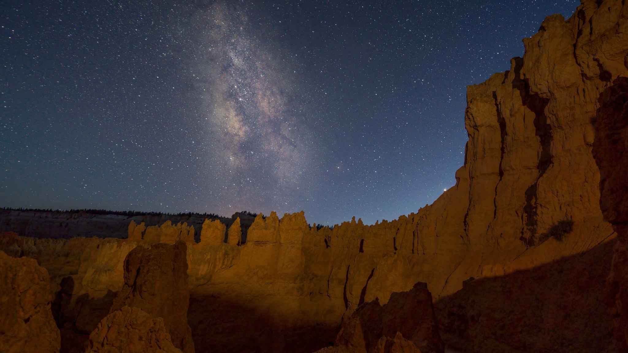 Rocky canyon cliffs under a clear night sky with the Milky Way galaxy visible above, illuminated by soft natural light.