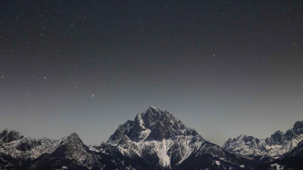 Snowy mountain peaks under a clear night sky filled with stars. The tallest peak is centered, with smaller peaks on either side, all in soft, dim lighting.