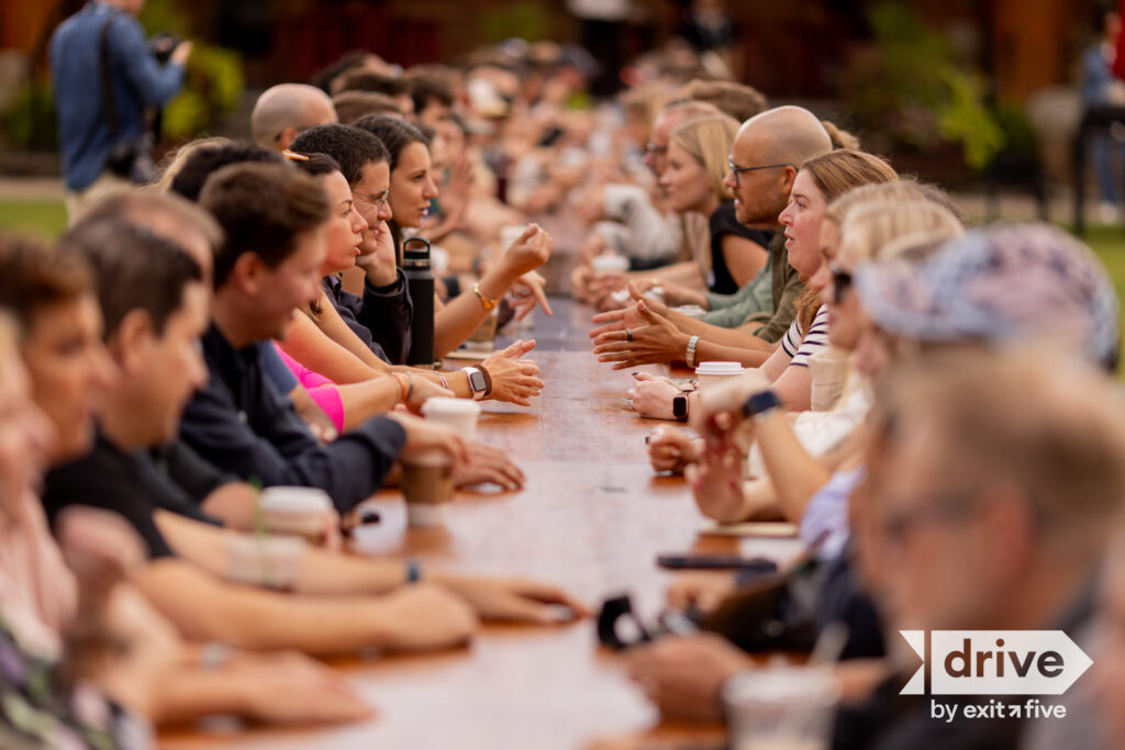 A large group of people sit on both sides of a long wooden table outdoors, talking and interacting. The focus is on the middle of the group. A drive by exit five logo appears in the bottom right corner.