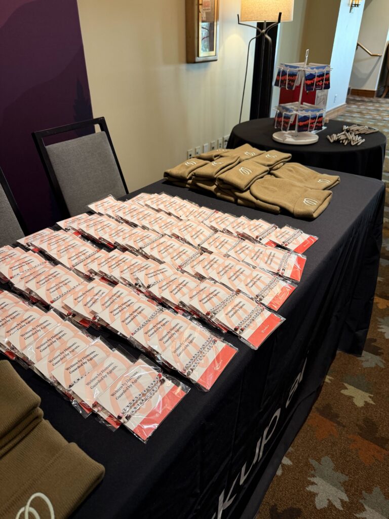 A table covered with rows of name badges, tan beanies, and a tiered stand holding lanyards creates an inviting drive conference registration area. A chair and a floor lamp are nearby, completing the welcoming setup for event attendees.