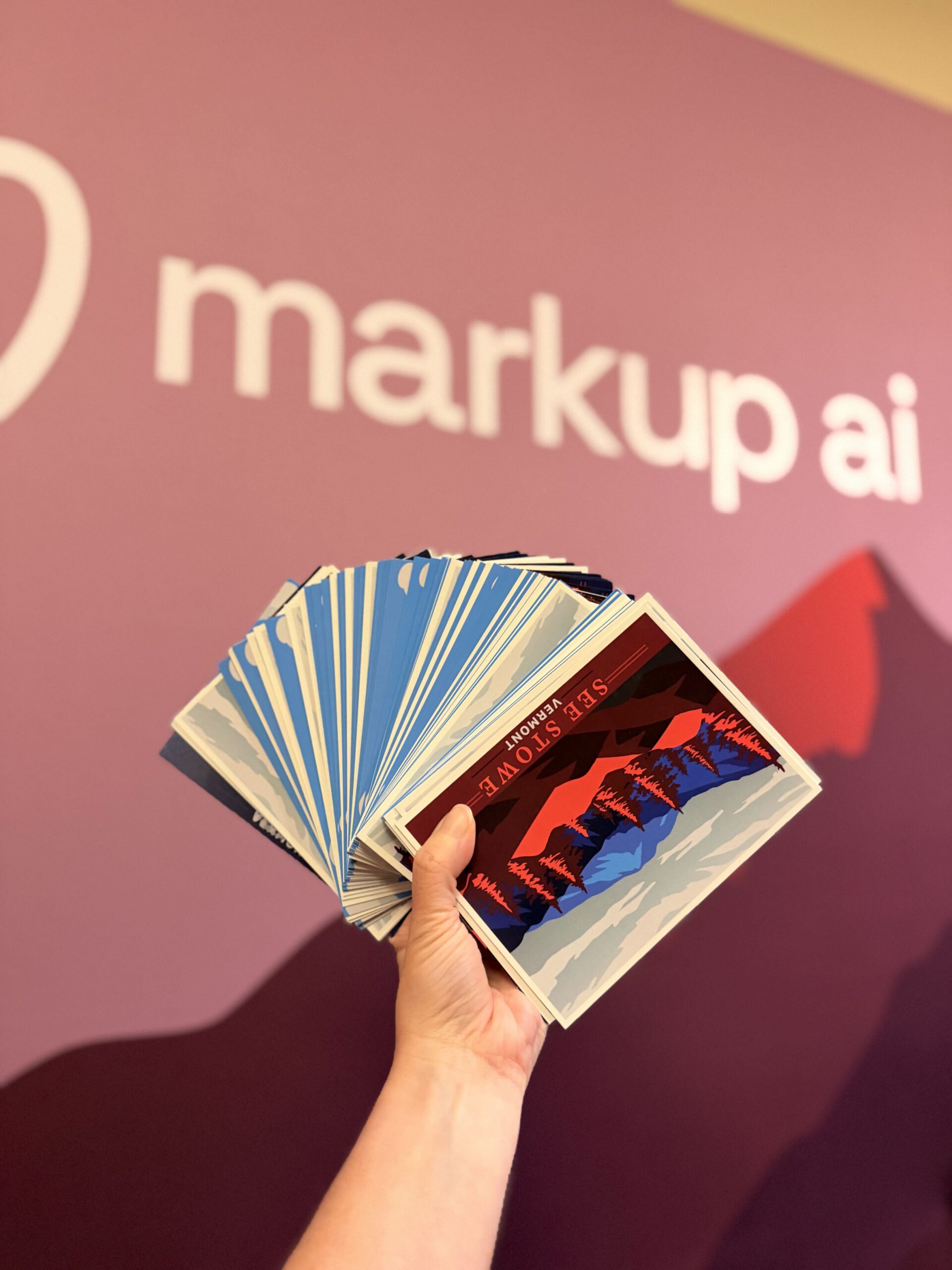 A hand holds a fanned-out stack of colorful cards in front of a purple wall with the words markup ai and a heart-shaped logo in white text, capturing the vibrant energy of the drive conference.