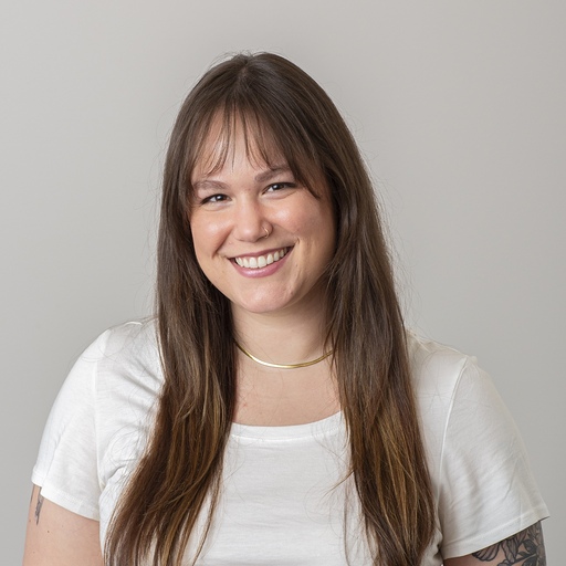 A young woman with long brown hair and bangs smiles at the camera. She is wearing a white t-shirt and a thin necklace, and has a nose ring. The background is plain and light gray.
