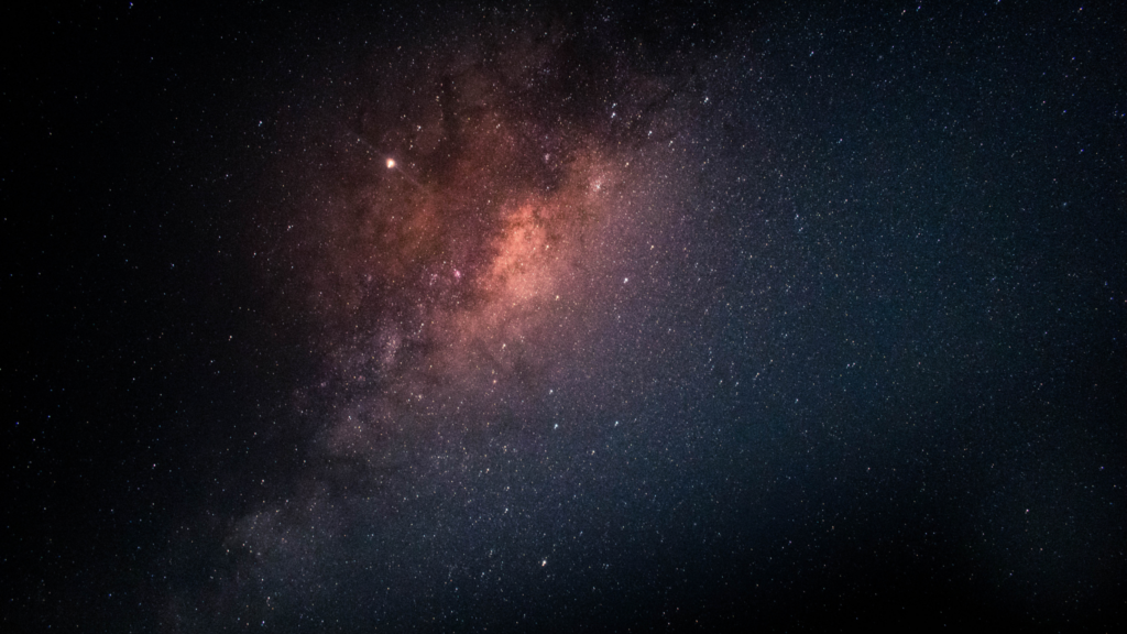 A breathtaking view of the Milky Way galaxy set against a deep black night sky, showcasing a dense, glowing band of stars, colorful interstellar dust clouds, and countless sparkling stars extending into the vast darkness of space.