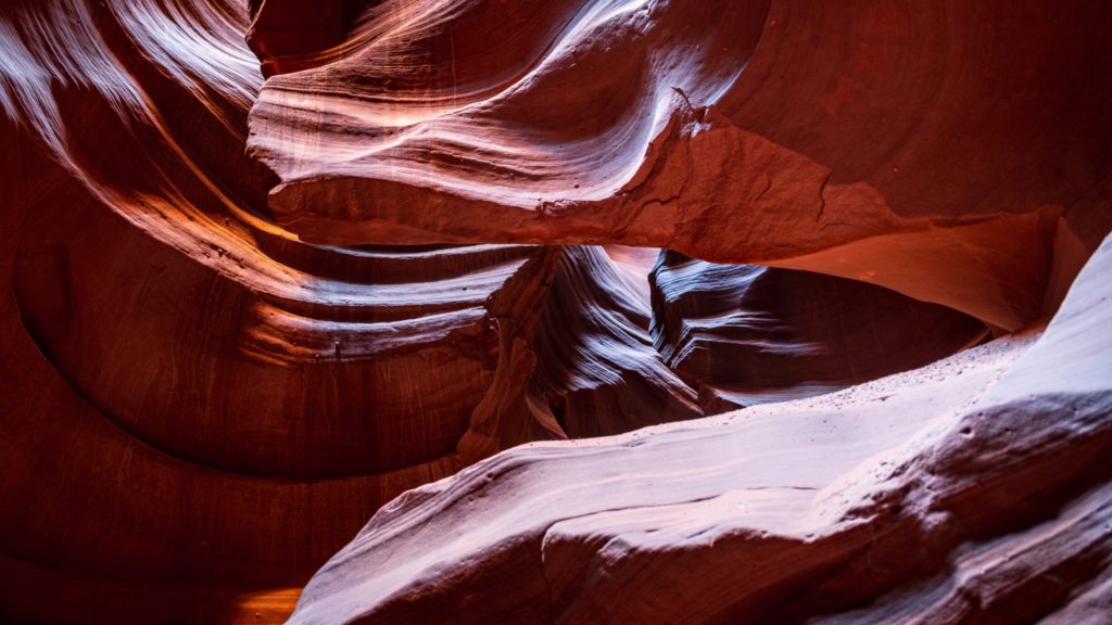 Sunlight illuminates the flowing, sculpted red sandstone walls of a slot canyon, highlighting the smooth curves and textures of the rock formations.