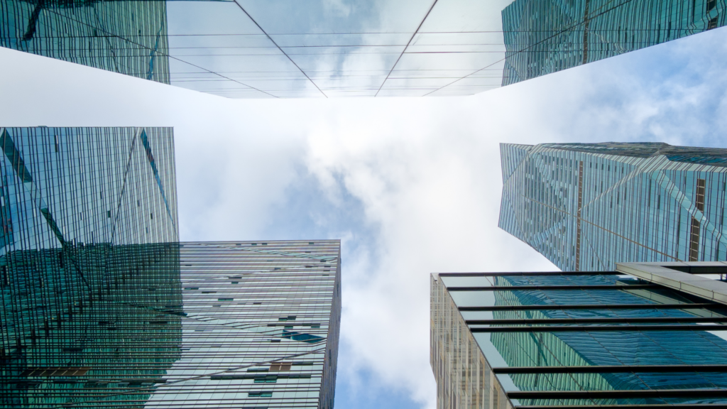 Four tall glass skyscrapers viewed from below, reflecting the sky and clouds above, converge to form a geometric frame of blue sky in the center of the image.