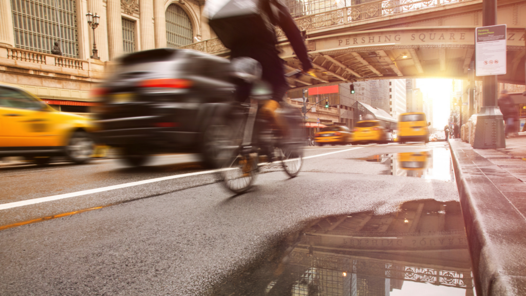 A cyclist rides through a rain-slicked New York City street near Pershing Square, with blurred cars and yellow taxis passing by.