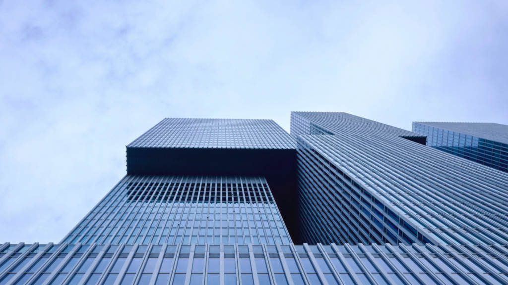 Looking up at modern skyscrapers with glass facades against a blue sky, the buildings feature sharp lines and angles, creating a futuristic urban appearance.